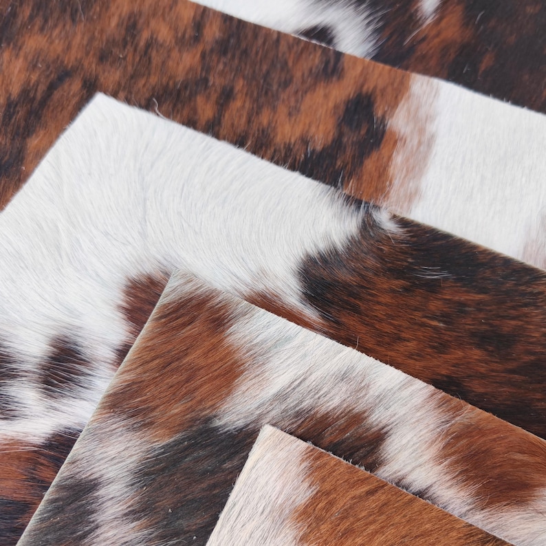 May include: Close-up of several pieces of cowhide leather. The leather has a natural, mottled pattern in shades of brown, white, and black. The texture appears soft and slightly raised, suggesting a high-quality material.
