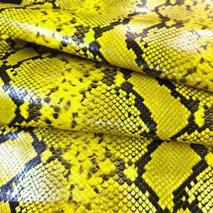 May include: A sheet of textured material featuring a vibrant yellow and black snakeskin pattern. The surface displays a repeating scale-like design with dark, irregular geometric markings layered over a bright, saturated yellow base color.