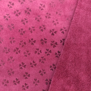 May include: Close-up of a magenta fabric with a repeating floral pattern. The fabric is a deep pink color with a soft, velvety texture. The floral pattern is a darker shade of pink, creating a subtle contrast. The fabric is folded over, revealing a solid magenta surface.