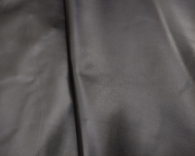 Op de afbeelding: Close-up van een zwarte lederen stof met een gladde, licht glanzende textuur. De stof is dubbelgevouwen, waardoor een vouw in het midden ontstaat.