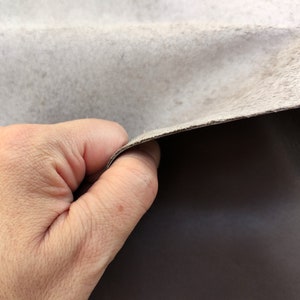 May include: A close-up of a hand holding a piece of light gray leather. The leather is folded over, revealing a darker gray underside.