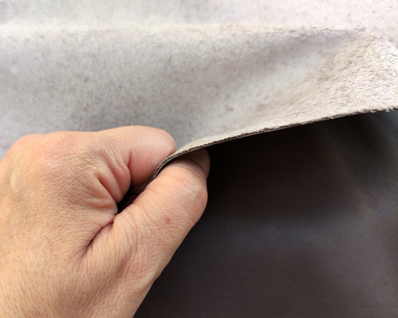 May include: A close-up of a hand holding a piece of light gray leather. The leather is folded over, revealing a darker gray underside.