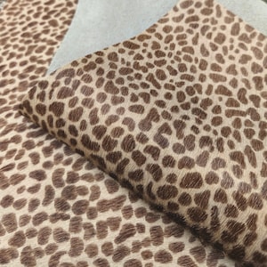 May include: Close-up of a folded piece of tan and brown animal print leather. The leather has a spotted pattern in varying shades of brown against a tan background. The texture appears soft and slightly raised.