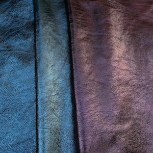 Pode incluir: Close-up de três secções de couro texturizado em tons de azul, verde-azulado e roxo. O couro tem um brilho metálico, com grão visível e variações subtis de cor na sua superfície. As secções são separadas por costuras escuras.