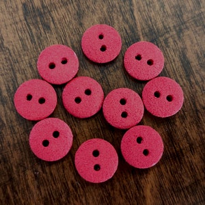Peut inclure: Dix boutons rouges avec deux trous chacun. Les boutons sont disposés en cercle sur une surface en bois.