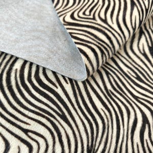 Può includere: Un pezzo di pelle animale con un motivo a strisce zebrate bianche e nere. La superficie superiore ha una finitura testurizzata con pelo, mentre il lato inferiore rivela un materiale grigio chiaro, liscio e scamosciato, esposto da un angolo ripiegato.