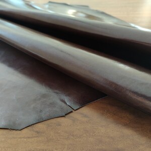 May include: A piece of dark brown leather with a glossy finish. The leather is folded in half and appears to be soft and supple.