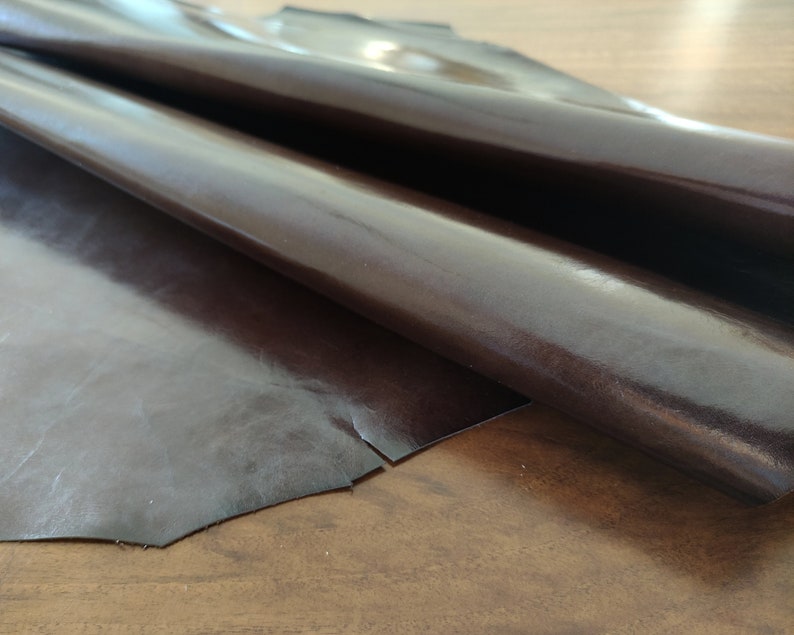 May include: A piece of dark brown leather with a glossy finish. The leather is folded in half and appears to be soft and supple.
