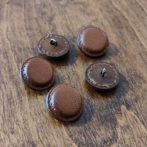 Peut inclure: Cinq boutons en cuir marron avec un bord cousu et une boucle métallique sur le dessus.