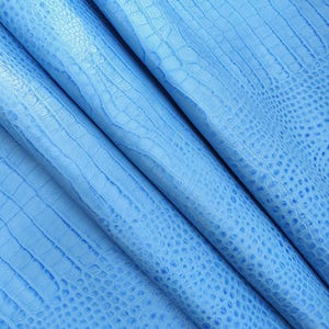 Puede incluir: Primer plano de cuero azul claro con textura de piel de cocodrilo. El cuero está doblado, revelando el patrón detallado y sutiles variaciones de color. El material tiene un acabado liso y ligeramente brillante, lo que sugiere alta calidad.