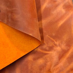 Peut inclure: Gros plan sur un morceau de cuir marron avec un coin plié, révélant un dessous orange vif. Le cuir présente une surface texturée avec des variations naturelles de couleur et de ton, suggérant un matériau de haute qualité. L'aspect général est rustique et chaleureux.