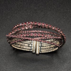 May include: A silver-toned bracelet with multiple bands of pink gemstones. The bracelet features a hinged clasp and a crisscross design, creating a layered effect. The gemstones are set closely together, giving the bracelet a sparkling appearance.