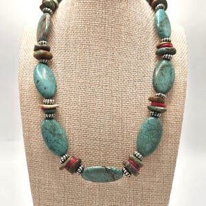 May include: A turquoise stone necklace with oval-shaped beads. The necklace features red, brown, and silver-colored accents. The necklace is displayed on a beige mannequin.