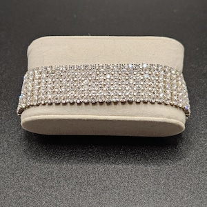 May include: A wide, silver-toned bracelet with multiple rows of sparkling rhinestones. The bracelet is displayed on a beige, rectangular jewelry display. The rhinestones catch the light, creating a dazzling effect. The bracelet appears to be made of metal.
