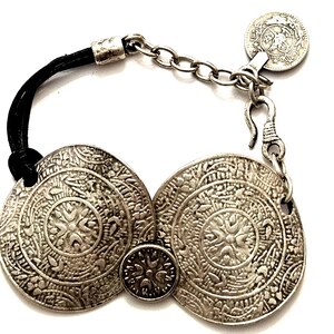 May include: Silver-toned bracelet featuring large, circular, engraved metal medallions. The bracelet has a black leather band and a chain with a coin-shaped charm. The medallions have intricate floral and geometric patterns. The bracelet is designed to be worn on the wrist.