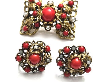 Puede incluir: Un conjunto de joyas vintage que incluye un broche y pendientes a juego. El conjunto presenta un diseño de filigrana de metal dorado, adornado con piedras de cabujón rojas y pequeños detalles rojos y blancos. El broche tiene forma de lazo.