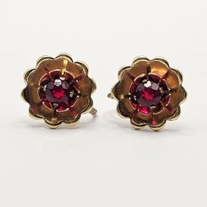 May include: A pair of vintage flower-shaped earrings. Each earring features a central, round, deep red gemstone surrounded by gold-toned petals. The petals have a reddish-brown interior and a scalloped edge. The earrings have a screw-back closure.