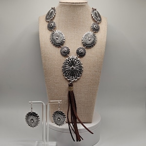 May include: A silver-coloured necklace and earring set displayed on a mannequin. The necklace features large, circular, sunburst-style pendants and a brown tassel. The matching earrings are also circular with a sunburst design. The jewellery is displayed on a beige fabric.