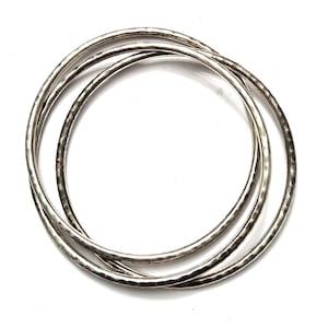 May include: Three silver bangles with a hammered texture are displayed against a white background. The bangles are circular and appear to be made of metal. The image is a top-down view of the jewelry.