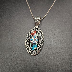 May include: Silver pendant necklace. The oval pendant features a floral mosaic design in red, blue, white, and green. The pendant has an ornate, openwork silver frame and hangs from a delicate silver chain.