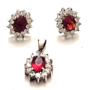 May include: A jewelry set featuring a pendant and matching earrings. Each piece has a central oval-shaped, deep red gemstone surrounded by a halo of smaller, clear gemstones. The metal setting appears to be silver-toned.