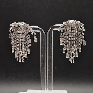 Puede incluir: Un par de pendientes colgantes plateados. Cada pendiente presenta un grupo de pequeños cristales redondos y transparentes en la parte superior, con múltiples hilos de cristales más pequeños en cascada hacia abajo. Los pendientes se exhiben sobre un soporte transparente sobre un fondo negro.