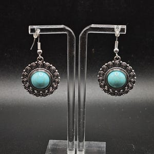 May include: A pair of silver-toned earrings with a turquoise-colored stone center. The earrings have a circular design with a spiral pattern around the stone. The earrings are displayed on a clear stand against a dark background.
