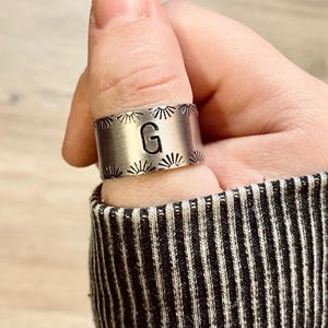 May include: A silver thumb ring with a sunburst design and the letter "G" engraved in the center.