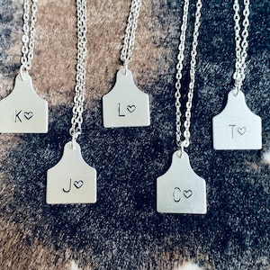 May include: Five silver necklaces with personalized silver tags shaped like cattle ear tags. Each tag has a letter and a heart engraved on it. The letters are K, L, J, C, and T.