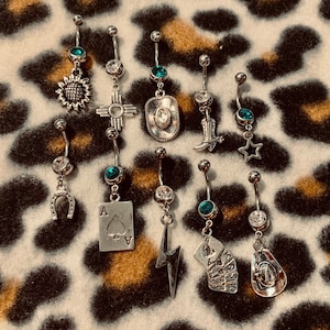 May include: A collection of nine silver belly button rings with various charms, including a horseshoe, a playing card, a lightning bolt, a cowboy hat, a boot, a star, a sunflower, a Native American symbol, and a playing card with multiple suits. The charms are all silver and have a turquoise gem at the top.