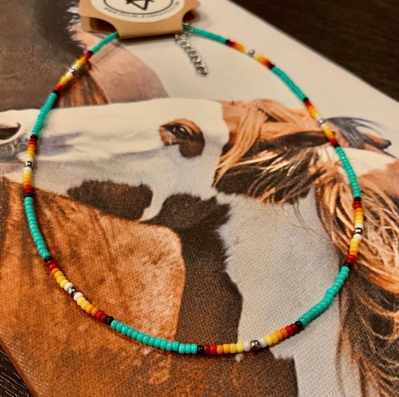 May include: A turquoise beaded necklace with red, yellow, white, and black accents. The necklace is on a fabric background with a horse pattern.