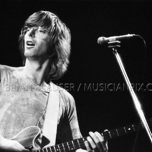 May include: Black and white photograph of a musician playing an electric guitar on stage. The musician is wearing glasses and a t-shirt. A microphone stand is in the background. The image is from BRIAN BLAUSER / MUSICIANPIX.COM.