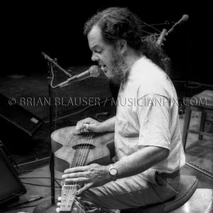 Peut inclure: Image en noir et blanc d'un musicien jouant d'une guitare acoustique. Le musicien est assis sur un tabouret avec une plaque d'immatriculation californienne qui dit "GIG RIG". Un microphone est positionné devant le musicien.