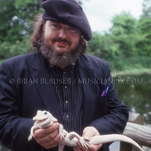May include: A man wearing a black beret and a black suit jacket holds a white alligator in his hands. The alligator is about 2 feet long and has its mouth open.