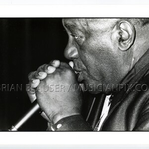 May include: Black and white photo of a man playing the harmonica. He is holding the harmonica to his mouth and blowing into it. The photo is taken from a low angle, so the man's face is the main focus of the image. The photo is credited to Brian Blauser/Musicianpix.com.