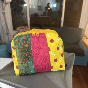 May include: A patchwork cosmetic bag with a yellow zipper. The bag features three vertical panels: green with orange spots, pink floral, and yellow with ladybugs and daisies. The bag is sitting on a gray and white striped surface.