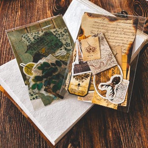 May include: A selection of vintage ephemera, including paper scraps, stickers, and botanical elements, presented in clear plastic sleeves. The items are displayed on a wooden surface, with a white book in the background.