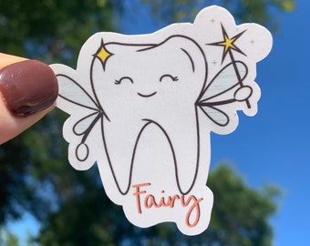 May include: A white tooth with a smiling face and fairy wings. The tooth is holding a magic wand with a yellow star on top. The text "Fairy" is written below the tooth in pink.