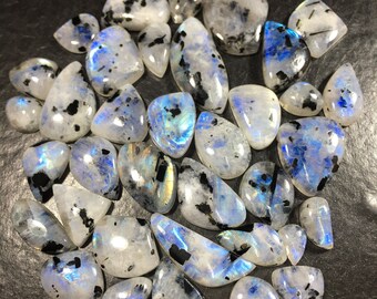 Puede incluir: Una colección de piedras preciosas de luna pulidas. Las piedras son principalmente blancas con inclusiones azules y negras, y varían en forma y tamaño. Algunas piedras tienen un brillo nacarado.