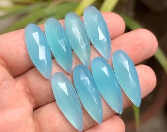 May include: A collection of eight faceted, teardrop-shaped gemstones in varying shades of light blue. The gems are held in a hand, with a blurred green background.