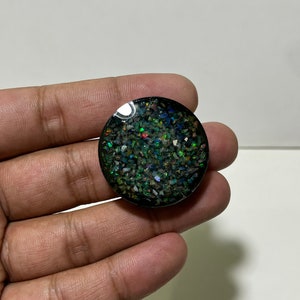 May include: A round, black object with a shimmering, speckled surface. The surface is filled with small, iridescent fragments in shades of green, blue, and red. The object is held in a hand against a white background.