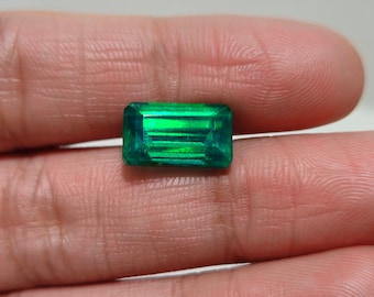 May include: A rectangular, emerald-cut gemstone with a vibrant green hue. The gem is held in a hand, showcasing its clarity and brilliance. The facets catch the light, creating a sparkling effect. This gemstone is likely for jewelry.