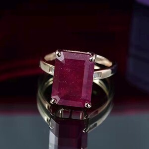 Puede incluir: Un anillo de oro con una gran gema rectangular de color rojo intenso. La gema es el punto focal, engastada en una banda de oro con un diseño simple y elegante. El anillo se exhibe sobre una superficie reflectante.