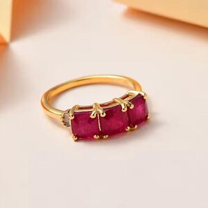 May include: A gold-toned ring featuring three rectangular, deep red gemstones set in a row. The ring has a simple band and decorative gold accents around the gemstones. The gemstones are a vibrant, saturated color, and the ring is set against a soft, neutral background.