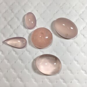 Könnte beinhalten: Fünf polierte Rosenquarz-Edelsteine in verschiedenen Formen: oval, rund, tropfenförmig und länglich. Die Steine sind zartrosa und wirken glatt und durchscheinend. Die Edelsteine werden auf einer weißen, strukturierten Oberfläche präsentiert.