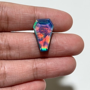 May include: A coffin-shaped gemstone with iridescent colors of blue, green, orange, and red. The gemstone is held between two fingers against a white background. The gem's facets reflect light, creating a shimmering effect.