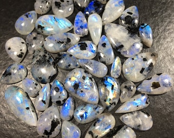 Puede incluir: Una colección de piedras lunares pulidas en varias formas, incluyendo lágrimas y óvalos. Las piedras muestran una base blanca lechosa con inclusiones azules y negras iridiscentes. Las piedras preciosas están dispuestas sobre una superficie oscura.