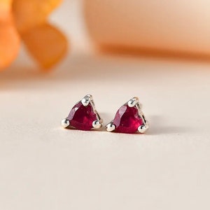 May include: A pair of silver stud earrings with triangular, deep red gemstones. Each earring has a silver setting with three small silver beads at the base of the triangle. The earrings are displayed on a light-colored surface.