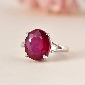 May include: A silver ring featuring a large, oval-cut ruby gemstone. The ruby is a deep red color and is held in place by four prongs. The ring's band splits into two delicate strands as it approaches the gemstone.