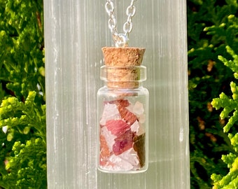 Puede incluir: Un collar de cadena de plata con un pequeño colgante de botella de vidrio lleno de cristales rosas y rojos. La botella tiene un tapón de corcho.
